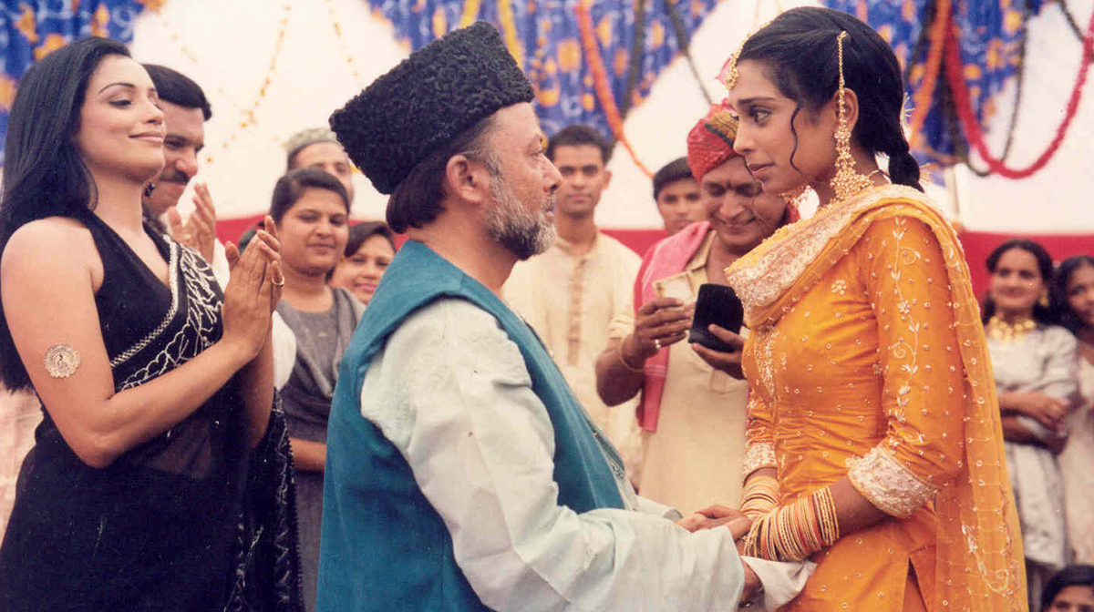 An elderly man with a grey beard holds hands with a young Indian woman in heavily embroidered clothing. They are surrounded by wedding guests.